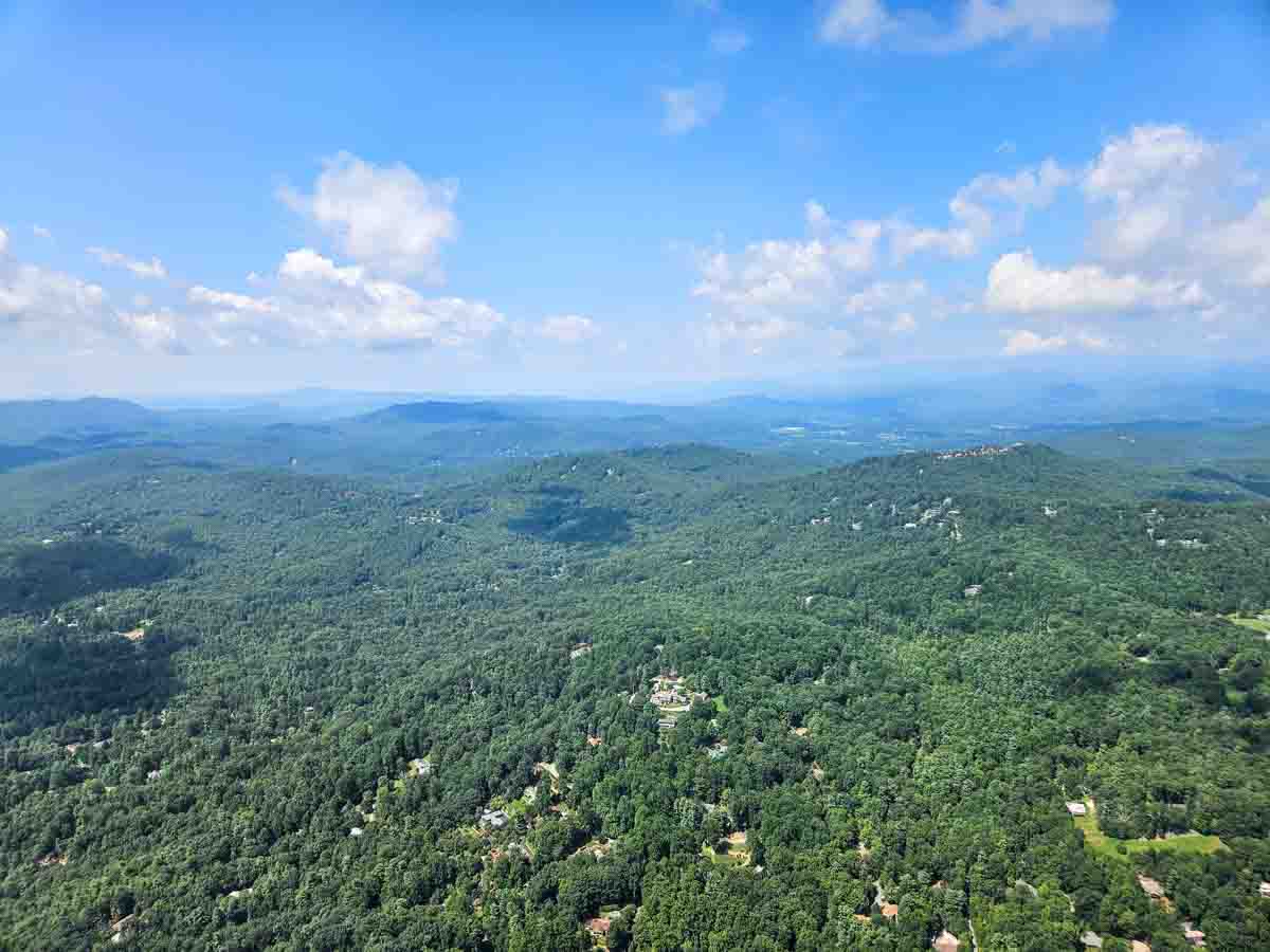 View of tree-covered hills with mountains in the background and a blue sky, taken from an airplane