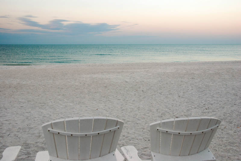 Backs of 2 Adirondack chairs on an empty beach, facing the ocean