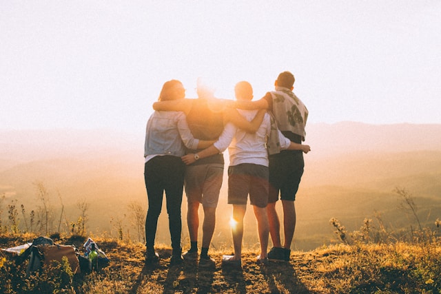 Backs of 4 people with arms around each other looking at the setting sun. Photo by Helena Lopes on Unsplash