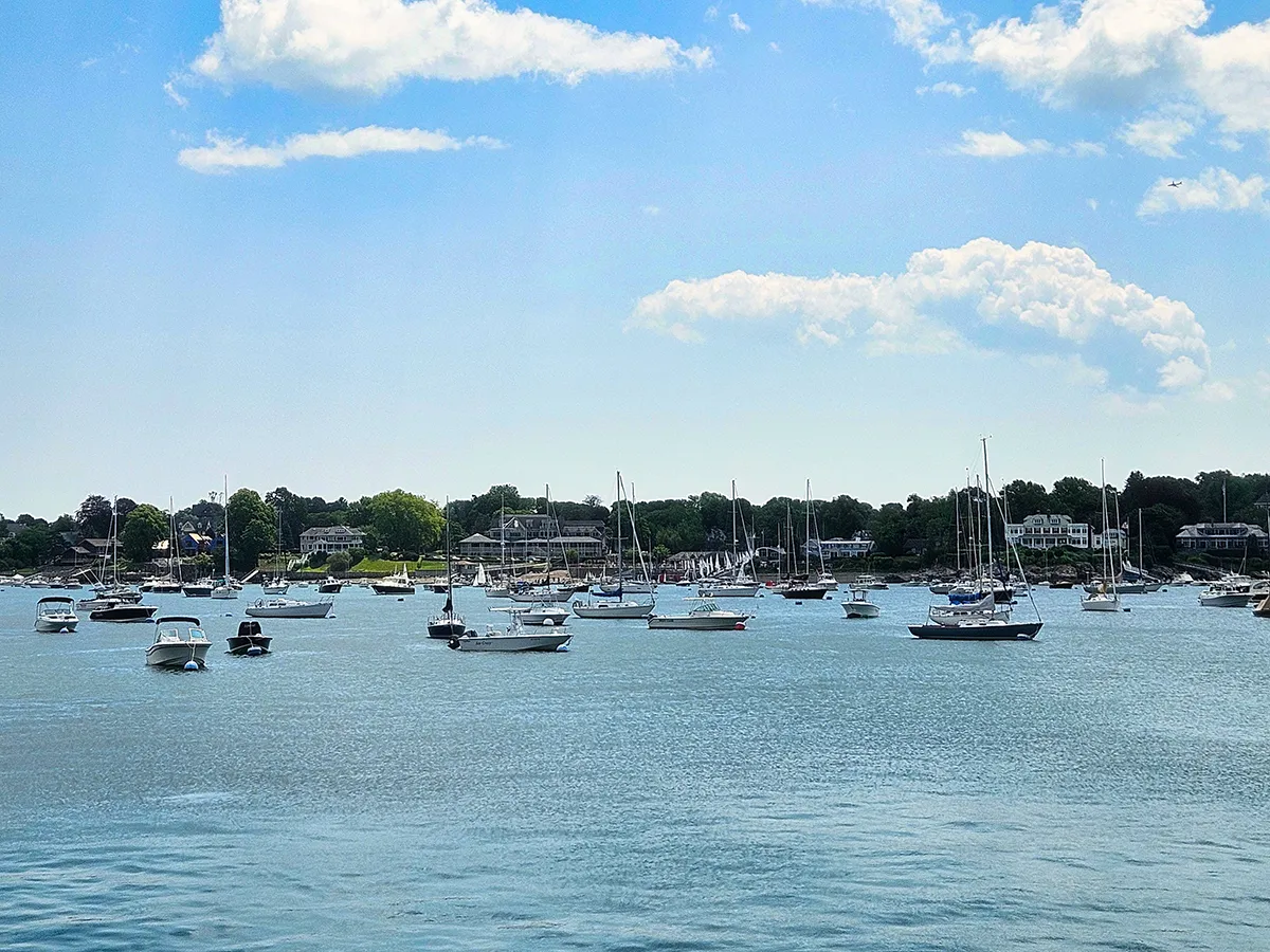 Photo of boats in a harbor, shore behind them, blue water, blue sky with white clouds